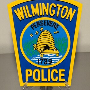 May include: A blue and yellow Wilmington Police badge. The badge features the words "Wilmington" and "Police" in yellow, with "Persevere" above a yellow beehive emblem. The year "1739" is also displayed.