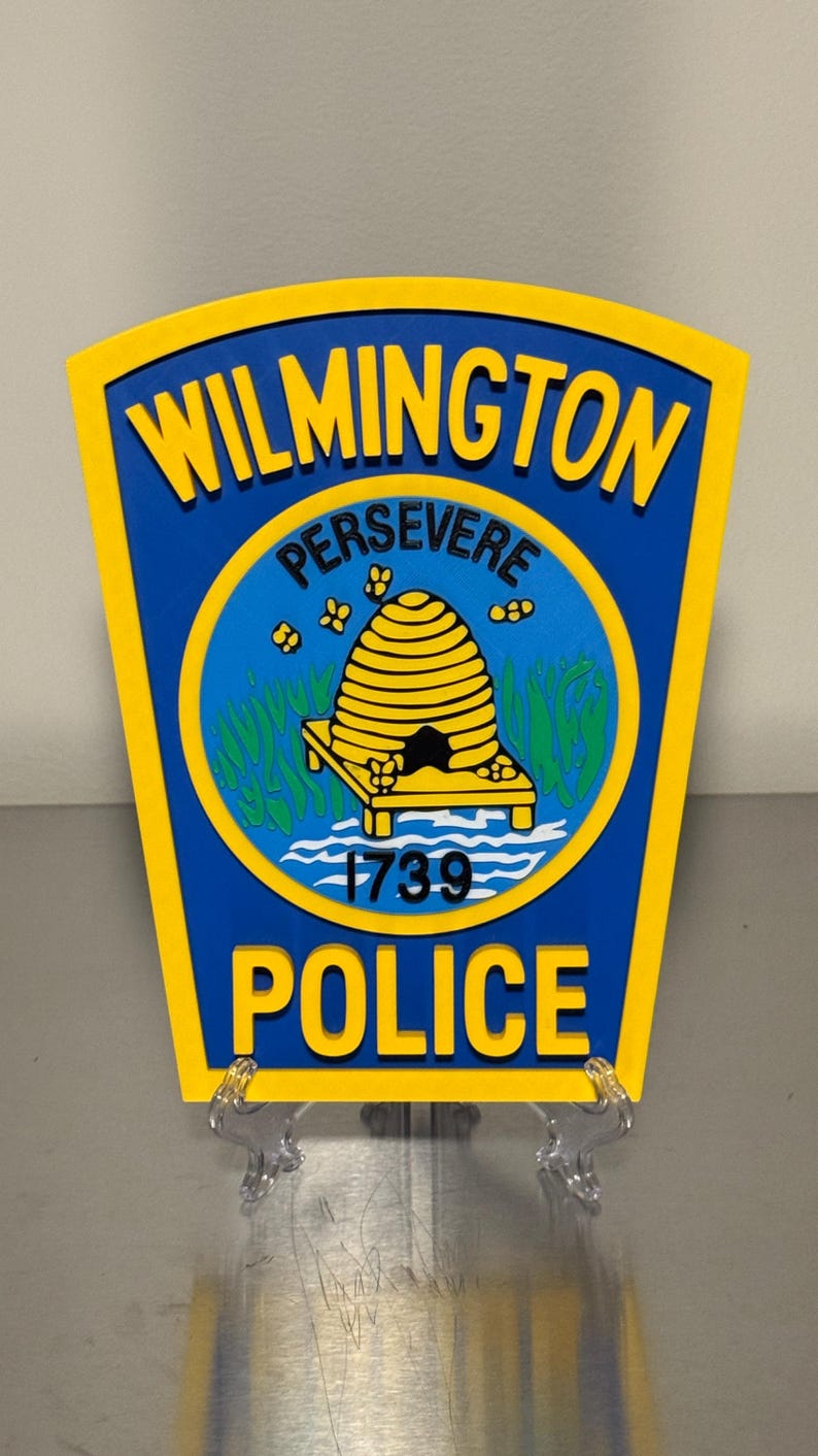 May include: A blue and yellow Wilmington Police badge. The badge features the words "Wilmington" and "Police" in yellow, with "Persevere" above a yellow beehive emblem. The year "1739" is also displayed.