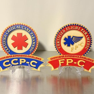 May include: Two circular plaques featuring the text Certified Critical Care Paramedic with a red star of life and Certified Flight Paramedic with a blue star and wing. Below each are banners reading Knowledge.Experience.Excellence with CCP-C and FP-C.
