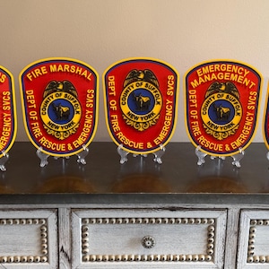 May include: Five red, yellow, and blue emergency service badges. Each badge has a unique title: Communications, Fire Marshal, Emergency Management, and Emergency Medical Services. The badges are displayed on clear stands.