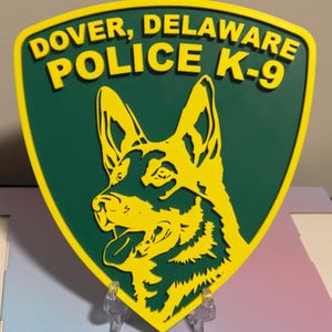 May include: A green and yellow shield-shaped sign with the text "Dover, Delaware Police K-9" and a silhouette of a German Shepherd dog's head.