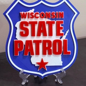 May include: A blue, white, and red shield-shaped sign with the words "WISCONSIN STATE PATROL" in red letters. The state's outline is in white, and a red star is at the bottom. The sign is displayed on a clear stand.