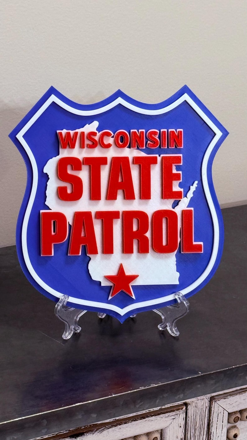 May include: A blue, white, and red shield-shaped sign with the words "WISCONSIN STATE PATROL" in red letters. The state's outline is in white, and a red star is at the bottom. The sign is displayed on a clear stand.