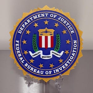 May include: A blue and gold Federal Bureau of Investigation (FBI) seal. The seal features the text "Federal Bureau of Investigation" and "Department of Justice". The center emblem includes the words "Fidelity, Bravery, Integrity".
