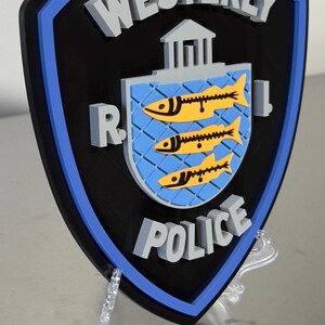 May include: A black, blue, and gray Westerly Police badge. The badge features the words "Westerly Police" in gray, with a blue and yellow emblem of three fish. The badge is displayed on a clear stand.