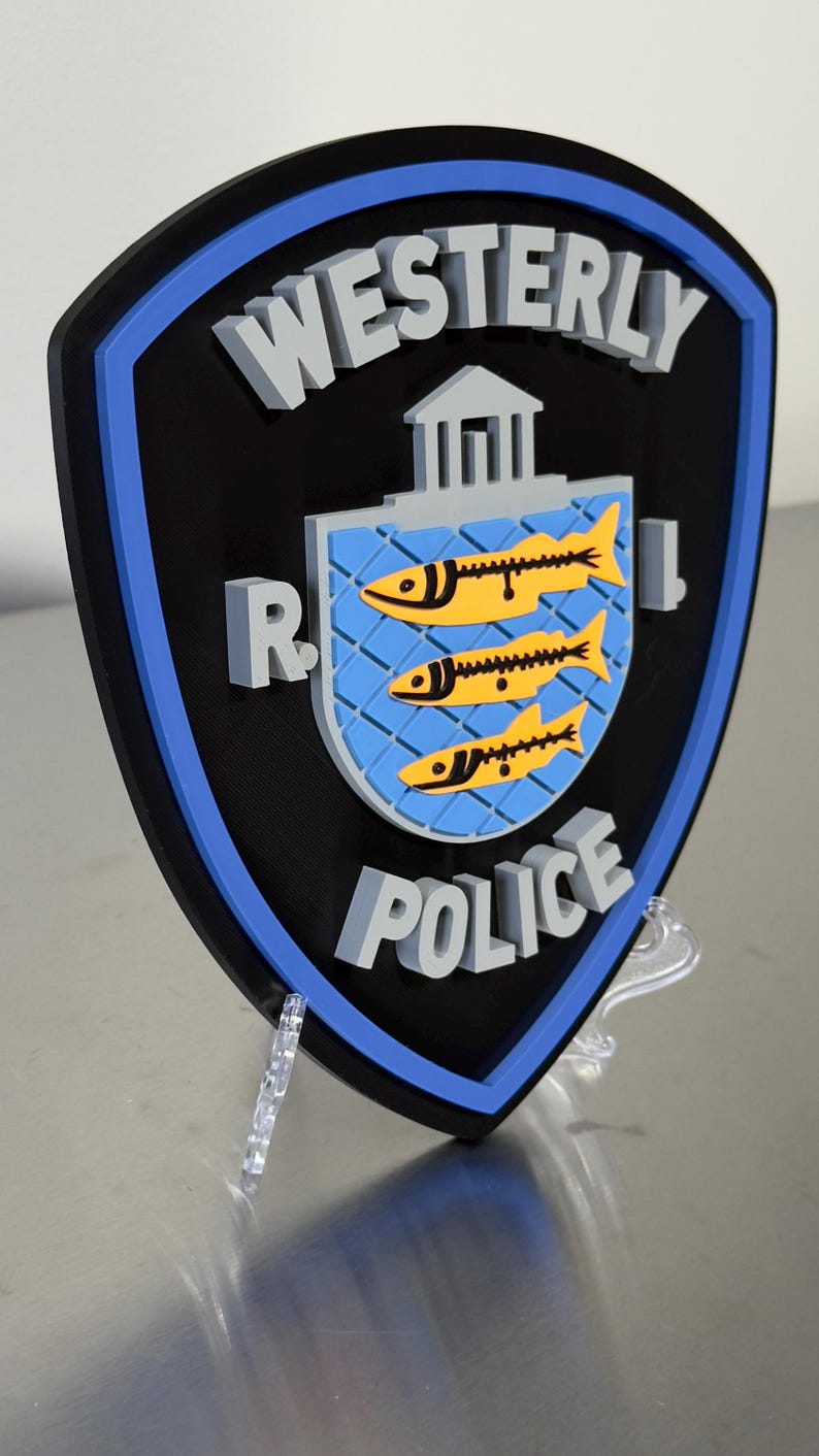 May include: A black, blue, and gray Westerly Police badge. The badge features the words "Westerly Police" in gray, with a blue and yellow emblem of three fish. The badge is displayed on a clear stand.