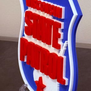 May include: A 3D-printed sign featuring the words "Wisconsin State Patrol" in red, white, and blue. The sign is shaped like a shield with a white outline and a red star. The sign is displayed on a clear stand.