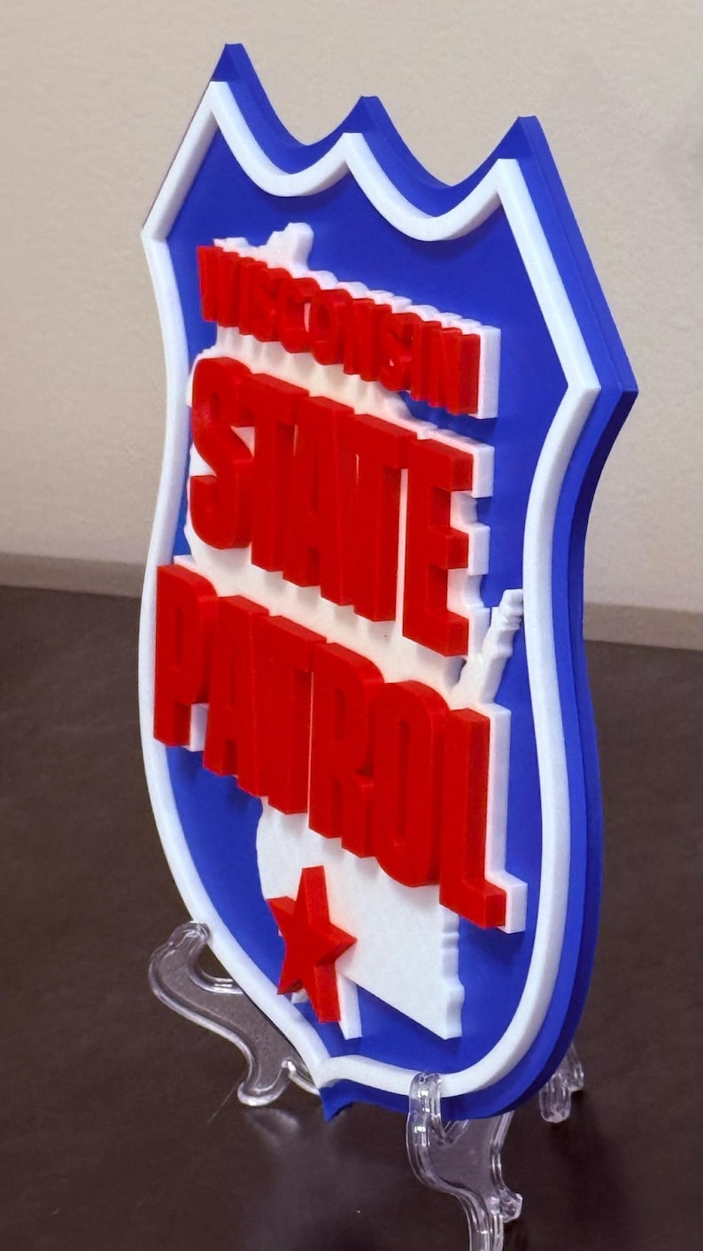 May include: A 3D-printed sign featuring the words "Wisconsin State Patrol" in red, white, and blue. The sign is shaped like a shield with a white outline and a red star. The sign is displayed on a clear stand.