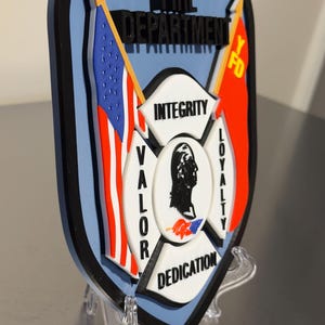 Op de afbeelding: Een schildvormig embleem voor de brandweer van Yonkers, met de Amerikaanse vlag en een rode vlag met "YFD". Het embleem bevat de woorden "Integriteit", "Moed", "Loyaliteit" en "Toewijding". Het embleem wordt op een transparante standaard getoond.