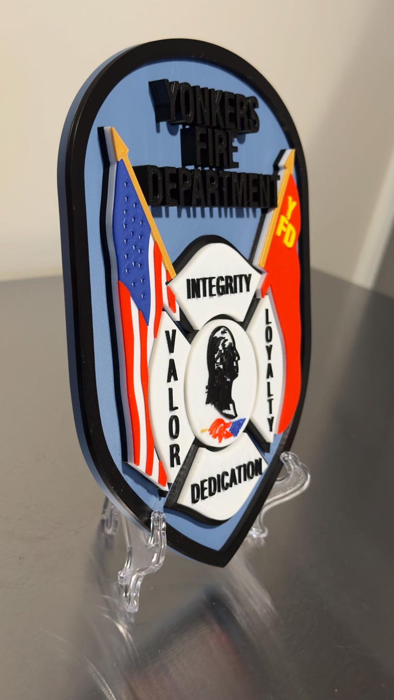 Op de afbeelding: Een schildvormig embleem voor de brandweer van Yonkers, met de Amerikaanse vlag en een rode vlag met "YFD". Het embleem bevat de woorden "Integriteit", "Moed", "Loyaliteit" en "Toewijding". Het embleem wordt op een transparante standaard getoond.