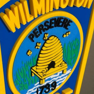 May include: A blue and yellow Wilmington Police Department emblem. The emblem features the word "Wilmington" in yellow, and the word "Police" in yellow. The center has a blue background with a yellow beehive and the word "Persevere".