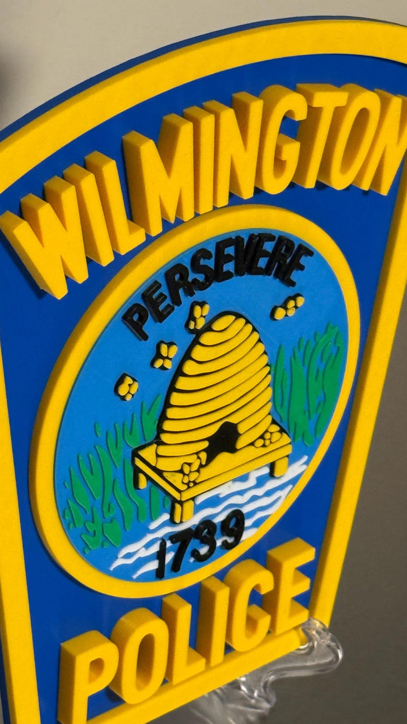 May include: A blue and yellow Wilmington Police Department emblem. The emblem features the word "Wilmington" in yellow, and the word "Police" in yellow. The center has a blue background with a yellow beehive and the word "Persevere".