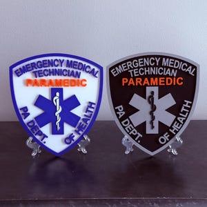 May include: Two shield-shaped plaques featuring the text EMERGENCY MEDICAL TECHNICIAN PARAMEDIC PA DEPT. OF HEALTH and a Star of Life symbol. One plaque has a white background with blue text and accents, while the other has a black background with silver text.