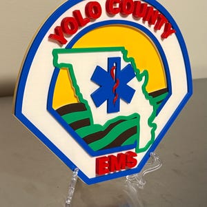 May include: A multi-layered sign featuring the words "YOLO COUNTY EMS" in red, with a blue and white outline. The centre includes a blue star of life symbol and a green outline of a state. The background is yellow.