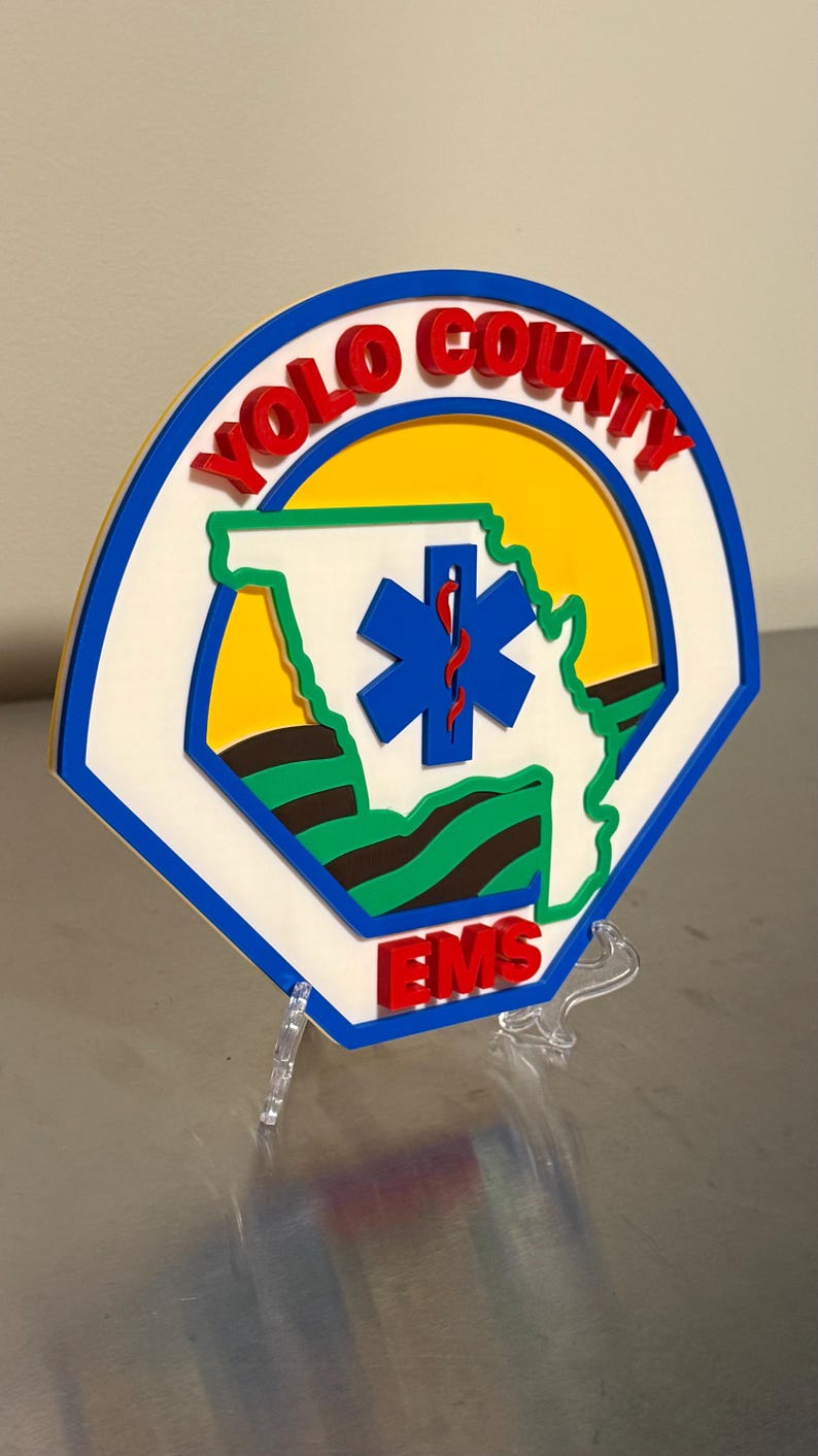May include: A multi-layered sign featuring the words "YOLO COUNTY EMS" in red, with a blue and white outline. The centre includes a blue star of life symbol and a green outline of a state. The background is yellow.