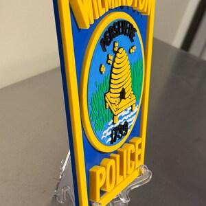 May include: A blue and yellow Wilmington Police badge with the words "Wilmington" and "Police" in yellow. The badge features a beehive and the year 1739. The badge is displayed on a clear plastic stand.