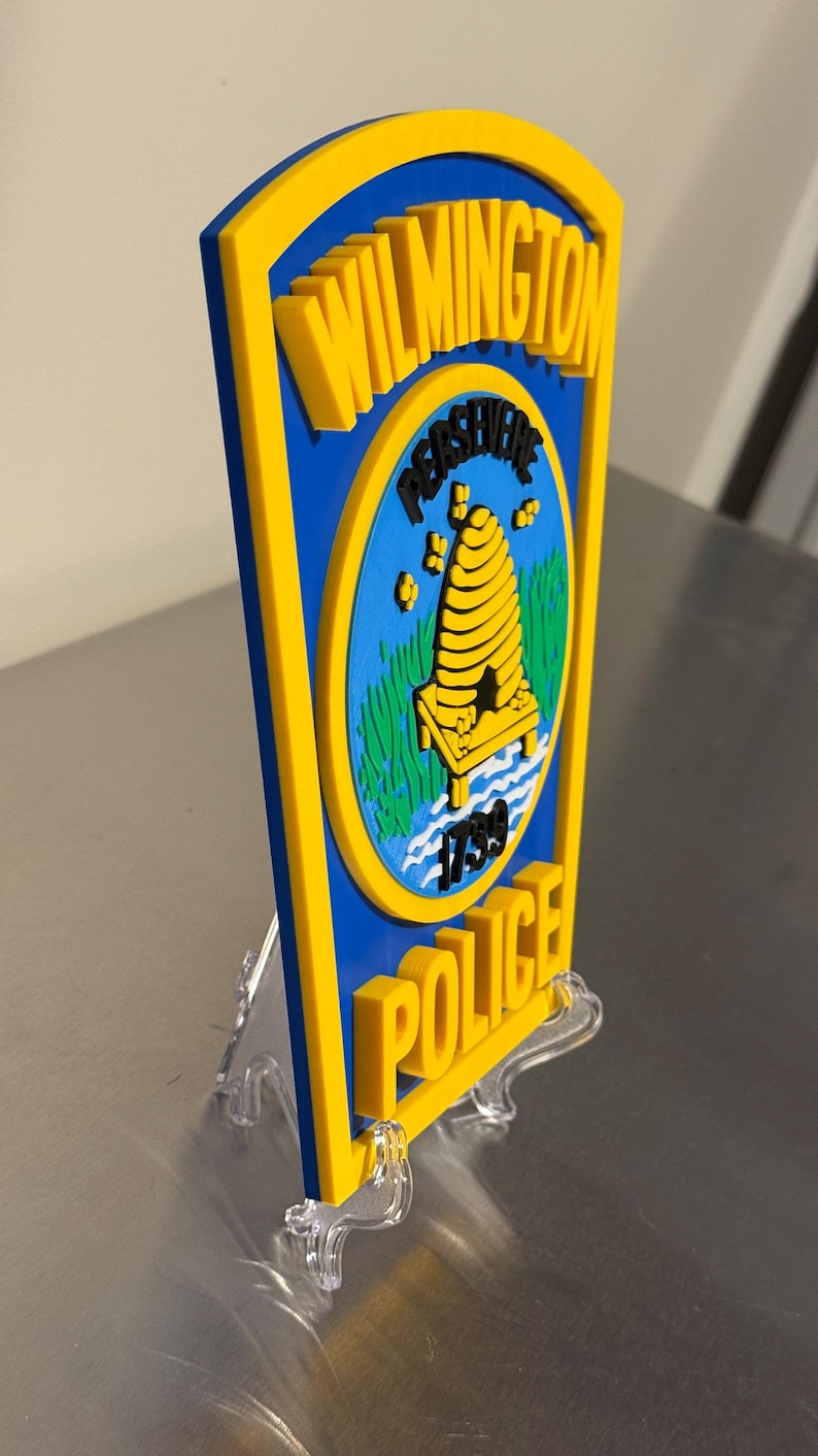 May include: A blue and yellow Wilmington Police badge with the words "Wilmington" and "Police" in yellow. The badge features a beehive and the year 1739. The badge is displayed on a clear plastic stand.