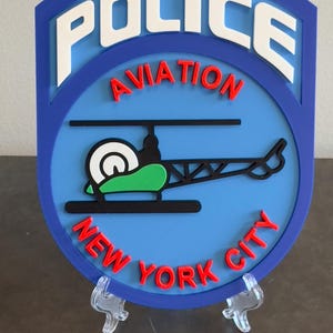 May include: A blue and white police aviation badge with the words "POLICE AVIATION NEW YORK CITY" in red and white lettering. A black helicopter with a green and white rotor is in the center of the badge.