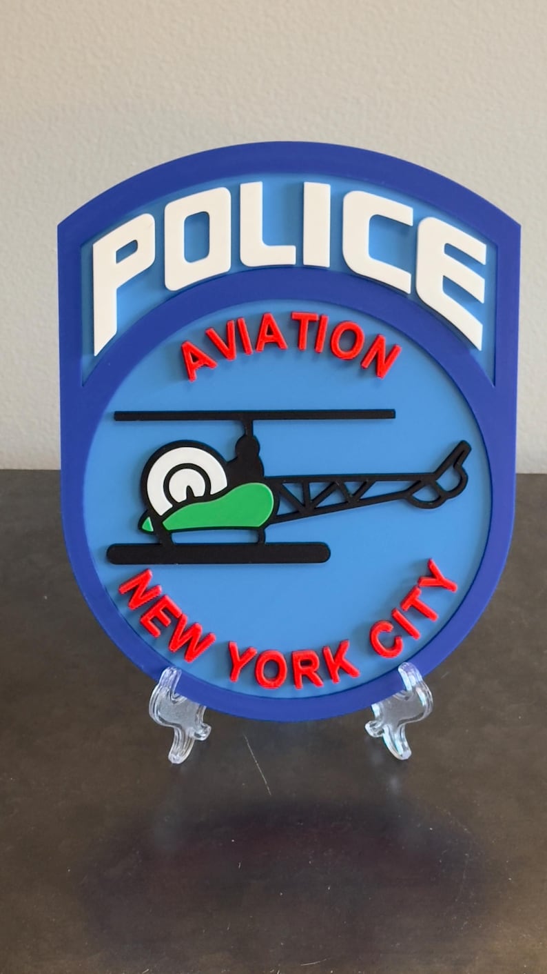 May include: A blue and white police aviation badge with the words "POLICE AVIATION NEW YORK CITY" in red and white lettering. A black helicopter with a green and white rotor is in the center of the badge.