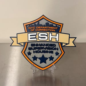 May include: A shield-shaped emblem with the text "NYC DEPARTMENT OF CORRECTION" above a banner that reads "ESH". Below, the text "ENHANCED SUPERVISION HOUSING" is displayed, with three dark blue stars. The emblem has a gray, orange, and dark blue color scheme.