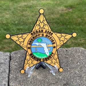 May include: A gold and black sheriff's badge in a star shape, featuring the text "Regan 5309 Sheriff's Office Marion County" and a Florida map graphic. The badge is displayed on a clear stand against a green background.