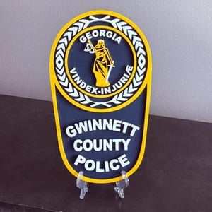 May include: A yellow and blue Gwinnett County Police badge with the words "Georgia", "Vindex-Injuria", "Gwinnett County", and "Police" in white lettering. The badge features a stylized image of a woman holding a scale and sword.