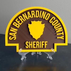 May include: A semi-circular sign featuring the text SAN BERNARDINO COUNTY SHERIFF in raised yellow lettering against a brown background. A yellow shield emblem is centered in the middle of the sign, which is supported by a clear plastic stand.