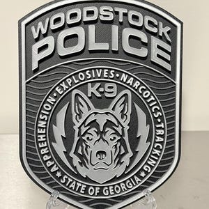 May include: A black and silver Woodstock Police badge with the words "WOODSTOCK POLICE" at the top. The badge features a K-9 unit emblem with the text "APPREHENSION EXPLOSIVES NARCOTICS TRACKING" and "STATE OF GEORGIA".