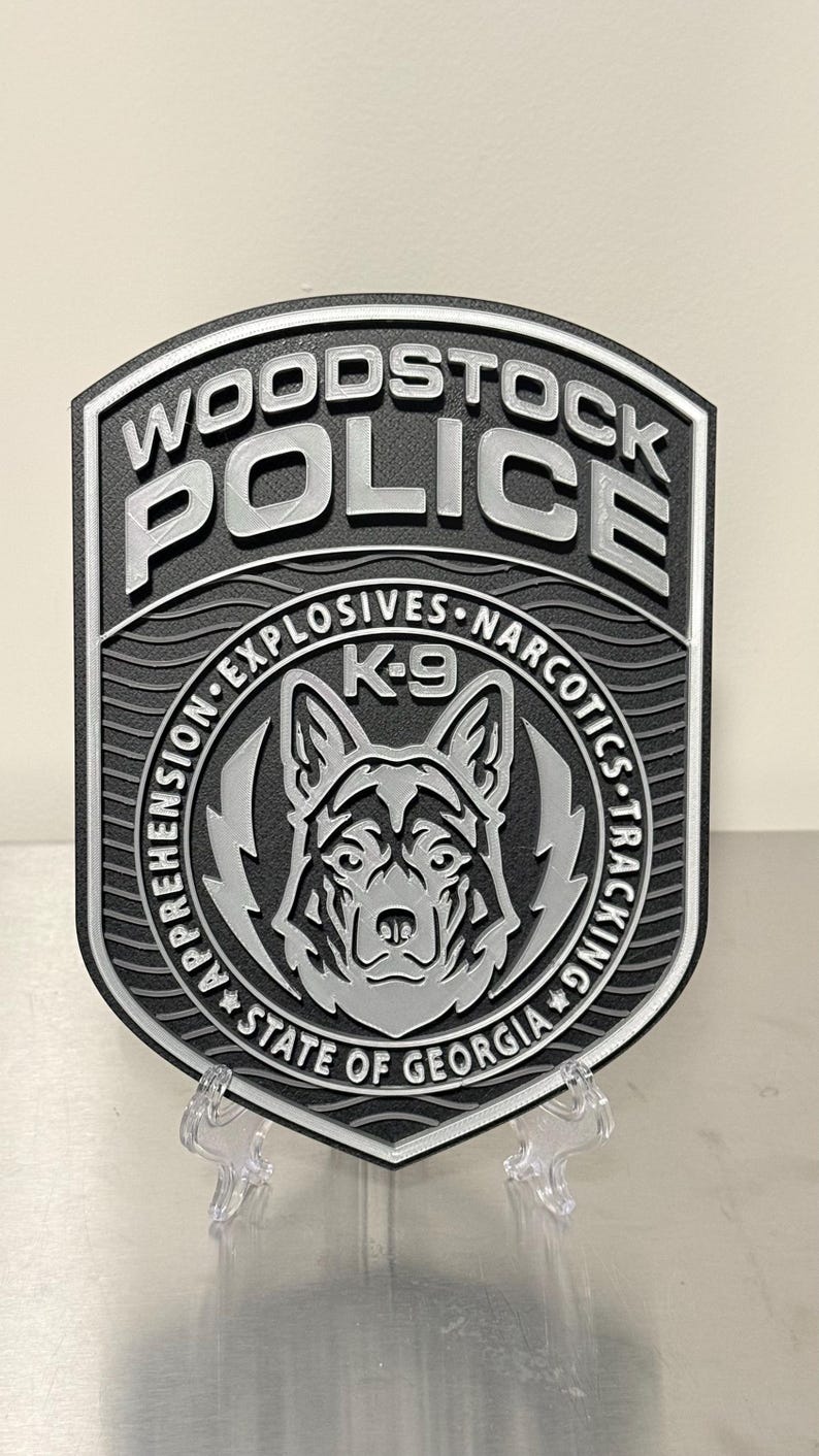May include: A black and silver Woodstock Police badge with the words "WOODSTOCK POLICE" at the top. The badge features a K-9 unit emblem with the text "APPREHENSION EXPLOSIVES NARCOTICS TRACKING" and "STATE OF GEORGIA".