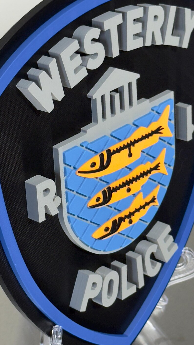 May include: A 3D sign featuring the words "WESTERLY POLICE" in gray block letters. The sign has a black background with a blue border. The center design includes a blue shield with three yellow fish.
