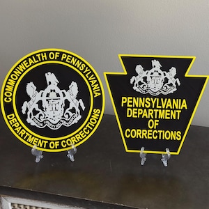 May include: Two Pennsylvania Department of Corrections signs. One is circular with yellow text and a black background, the other is a keystone shape. Both feature the state seal.