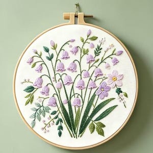 Könnte beinhalten: Ein runder Stickrahmen aus Holz mit weißem Stoff, der ein handgesticktes Blumenmotiv zeigt. Die Stickerei besteht aus zahlreichen hellvioletten glockenförmigen Blüten, grünen Blättern und Stängeln sowie zwei größeren hellvioletten Blüten mit gelben Mitten.
