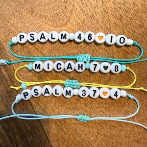 May include: Three adjustable cord bracelets featuring white round beads with black letters and numbers. The top bracelet reads PSALM 46:10 with an orange heart. The middle reads MICAH 7:8 with a green heart. The bottom reads PSALM 37:4 with an orange heart.