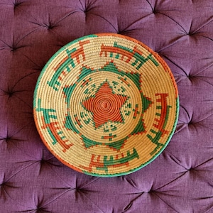 May include: A round, woven basket with a starburst design. The basket features a natural beige base with orange and green geometric patterns. The basket is approximately 12 inches in diameter and is placed on a purple, tufted surface.