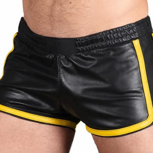 May include: Black shorts with a smooth, leather-like finish featuring a wide, elasticated waistband. The shorts are accented with bright yellow piping along the curved leg openings and side seams, creating a distinct contrast against the black.