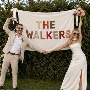 以下が含まれることがあります： THE WALKERSという文字がマルチカラーの柄入り布製大文字で記された、オフホワイトの長方形の布製バナー。バナーは白いリボンで吊るされており、右上隅には花のブーケが飾られている。