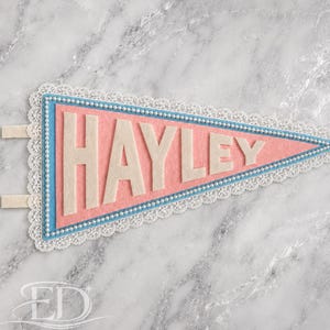 May include: A triangular felt pennant featuring the name HAYLEY in large cream-colored letters. The pennant has a pink center, a blue border, and is edged with white lace trim and a row of small white beads. Two cream felt tabs extend from the left.