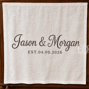 May include: A square off-white fabric napkin featuring dark brown embroidered text that reads Jason & Morgan with the date EST. 04.09.2026 centred below the names. The fabric has a visible woven texture and a stitched border along the edges.