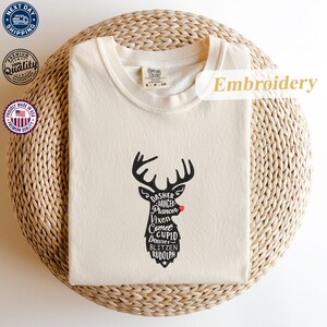 May include: Cream-colored t-shirt with a black reindeer design featuring the names of Santa's reindeer. The reindeer has a red nose. The shirt is folded on a woven surface. The word "Embroidery" is visible.