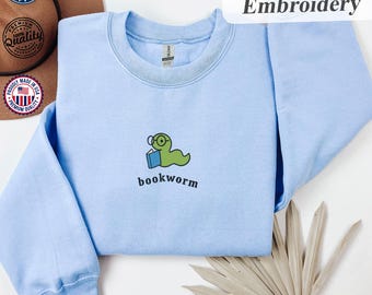 May include: Light blue sweatshirt with an embroidered design. The design features a green bookworm wearing glasses and holding a book, with the word "bookworm" below. The word "Embroidery" is also visible.