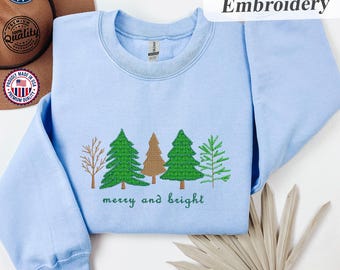 Può includere: Felpa azzurra con ricami di alberi di Natale e la scritta "merry and bright" in verde. Il design include vari stili di alberi in verde e marrone. La parola "Ricamo" è in un rettangolo bianco.