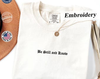 May include: Cream-colored t-shirt with the embroidered phrase "Be Still and Know" in black gothic lettering. A gold necklace with a pendant rests on the shirt. The word "Embroidery" is written in black. The shirt is labeled "Comfort Colors".