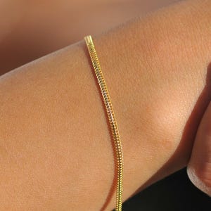 May include: A fine gold chain bracelet is worn on a wrist. The bracelet features a small charm. The person's hand is visible, with nails painted with a French manicure design. The bracelet is a simple, elegant piece of jewellery.