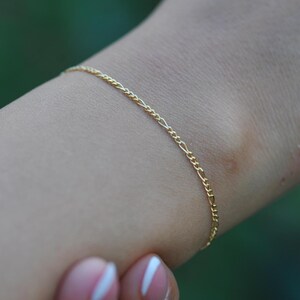 May include: A delicate gold Figaro chain bracelet. The bracelet is shown on a wrist, with a blurred green background. The bracelet's links alternate between two small and one elongated link.