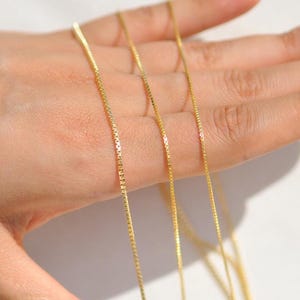 May include: Close-up of several fine gold box chain necklaces. The necklaces are held in a hand, highlighting their delicate design and the warm gold colour. The background is a soft, neutral tone, enhancing the necklaces' appearance.