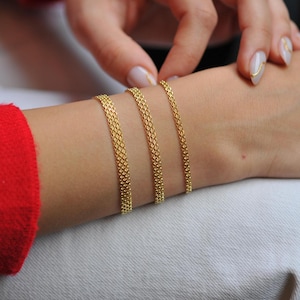 May include: Four gold chain bracelets of varying widths stacked on a person's wrist.
