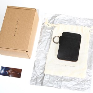 May include: A black leather key holder with a silver key ring. The key holder is packaged in a white drawstring bag and a brown cardboard box. The box has the text "ULAGOODS" printed on it.