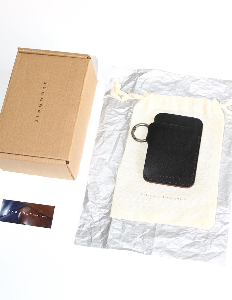 May include: A black leather key holder with a silver key ring. The key holder is packaged in a white drawstring bag and a brown cardboard box. The box has the text "ULAGOODS" printed on it.