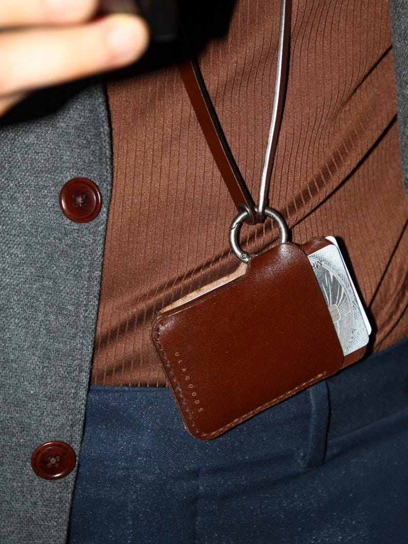May include: A brown leather card holder with a silver clasp and the text "ULAGGOODS" on the front. The card holder is attached to a brown and white lanyard.