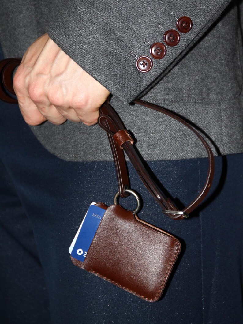 May include: A brown leather ID badge holder with a blue card inside. The holder is attached to a brown leather strap with a metal ring.